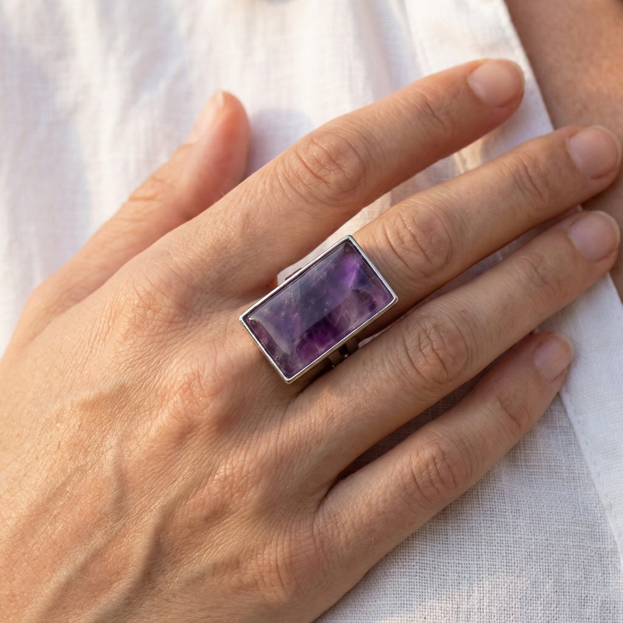 Prostokątny pierścionek z naturalnym fioletowym ametystem. Srebrna regulowana biżuteria na dłoni modelki.